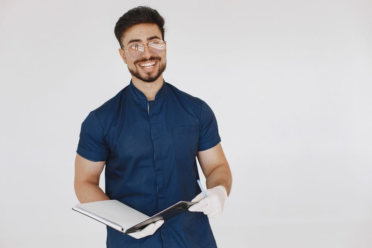 A cheerful medical professional in blue scrubs holding a notebook, wearing glasses and white gloves.