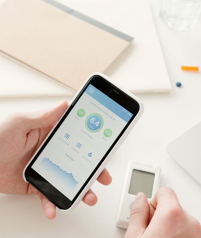Close-up of hands using a smartphone app to monitor glucose levels with a digital glucometer on a table.