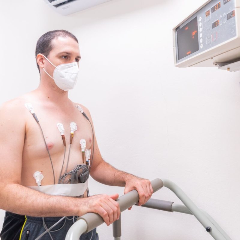 Man in medical exam wearing electrodes and face mask performing heart stress test on treadmill.